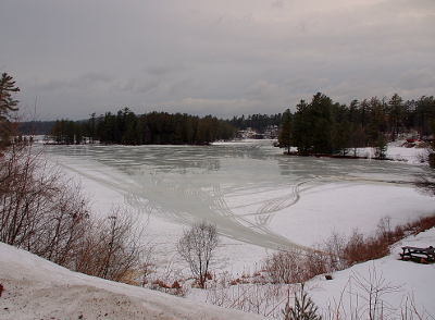 [The lake vees outward from the shore at this viewpoint. A snow-covered picnic table is at the right edge of the image. The water edges closest to shore are snowcovered except for the area where the snowmobiles have entered the lake when it was frozen. Evergreens are visible on the far shoreline.]